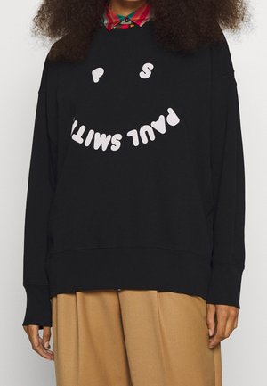 Personne portant un sweat-shirt noir avec un texte blanc incurvé formant "PAUL SMITH" qui crée un visage souriant, un pantalon beige et un col de chemise multicolore.
