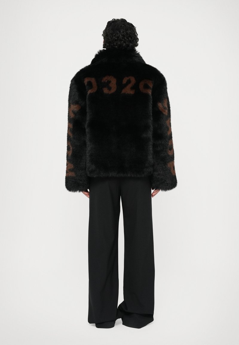 Sort faux fur jakke med store brune numeriske detaljer på ryggen og ærmerne, parret med sorte vide bukser.