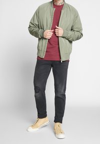 Olivinvihreä bomber-takki burgundinpunaisen collegepaidan päällä, mustutetuilla revityillä farkuilla ja beigeillä lenkkareilla, joissa on valkoiset pohjat.