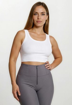 Nuori nainen, jolla on pitkät ruskeat hiukset, valkoinen lyhyt paita ja korkeavyötäröiset harmaat legginsit, joissa on etuvetoketju. Hän poseeraa yhdellä kädellä lanteilla.