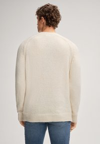 Uomo con capelli ricci che indossa un maglione lavorato a maglia color crema e jeans blu, in piedi di spalle contro uno sfondo semplice.