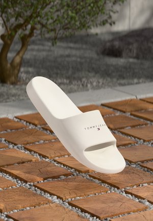 White slide sandal with an open toe, featuring a wide strap and embossed “TOMMY JEANS” logo. Smooth texture, minimalistic design, casual look.