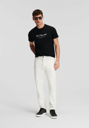 Black short-sleeve t-shirt with white logo, paired with white straight-leg jeans and black sneakers with white soles. Sunglasses complete the look.