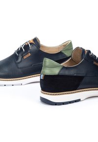 Chaussures en cuir noir avec un matériau texturé, accents en cuir vert, semelle blanche et couture marron. Dotées de lacets ronds et d'un tag de marque.