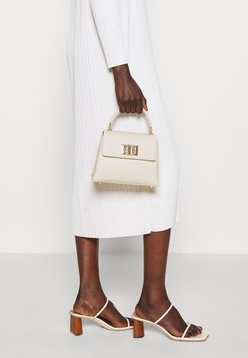 Jasbeżowa torebka w strukturalnym designie, z złotymi elementami i górnym uchwytem. Nosi ją do białej plisowanej sukienki i jasnych sandałów na obcasie.