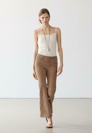 Vrouw loopt in beige spaghettibandtop, bruine suède uitlopende broek, zwarte sandalen en lange ketting tegen een effen grijze achtergrond.