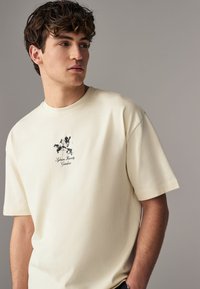 Cream-colored cotton T-shirt with short sleeves, featuring a graphic print of black flowers and text in the center. Smooth texture.