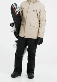 Veste beige isolante avec plusieurs poches, pantalon de neige noir et bottes de neige noires. Tenant un snowboard avec un design en noir et blanc.