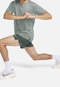 Camiseta atlética de color gris claro con un patrón, combinada con pantalones cortos de color verde oscuro. Modelo demostrando una zancada, vistiendo calcetas blancas y zapatillas.