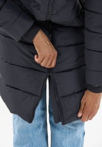 Svart puffjacka med kviltat mönster, dragkedja och ribbade mudd. Buren över blå denimjeans, med händer som justerar dragkedjan.