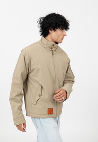 Beżowa wodoodporna kurtka z wysokim kołnierzem, zamkiem błyskawicznym z przodu oraz zatrzaskami. Posiada boczne kieszenie i naszywkę z logo na dolnym brzegu.