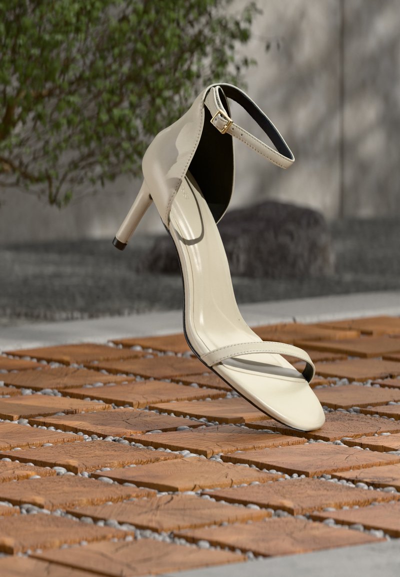 Sandale beige à talons hauts avec bride à la cheville posée sur des lattes en bois en extérieur, avec un arrière-plan flou de verdure et de béton.