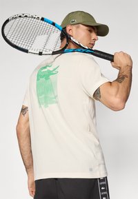 Kreem lühikeste varrukatega särk, mille seljal on roheline krokodilli graafika. Ingel hoiab sinist ja musta tennisereketit, kandes oliivirohelist mütsi.
