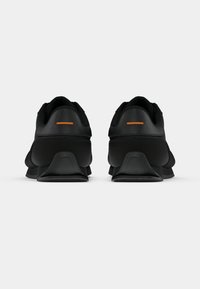 Ténis atléticos pretos com uma superfície lisa, apresentando uma faixa de destaque laranja no calcanhar e uma sola resistente e texturizada.