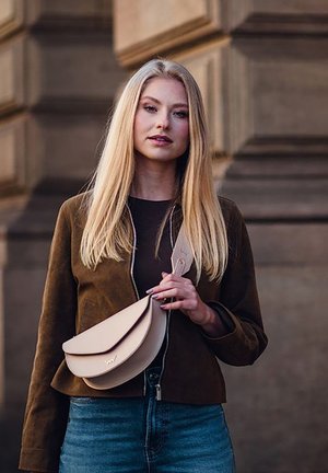 Junge Frau mit langen blonden Haaren trägt eine braune Jacke und blaue Jeans und hält eine beigefarbene Umhängetasche vor einem steinernen Hintergrund.
