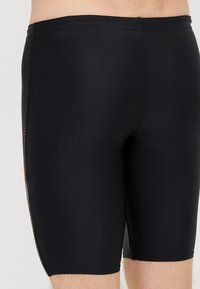 Μαύρο μαγιό σορτς με εφαρμοστή σχεδίαση, ελαστικό ύφασμα, μήκος έως το μεσαίο θηλυκό και πλευρική λεπτομέρεια με γεωμετρικό μοτίβο σε κόκκινες αποχρώσεις.