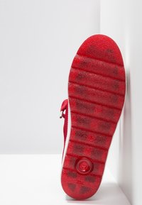 Chaussure en daim rouge avec une semelle en caoutchouc texturée, présentant des rainures horizontales et un accent circulaire. Fermeture éclair latérale sur la tige.