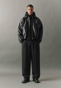 Giubbotto bomber in pelle nera con zip, colletto alto, abbinato a un maglione in maglia nera e pantaloni neri larghi. Scarpe nere spesse.