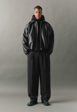 Giubbotto bomber in pelle nera con zip, colletto alto, abbinato a un maglione in maglia nera e pantaloni neri larghi. Scarpe nere spesse.