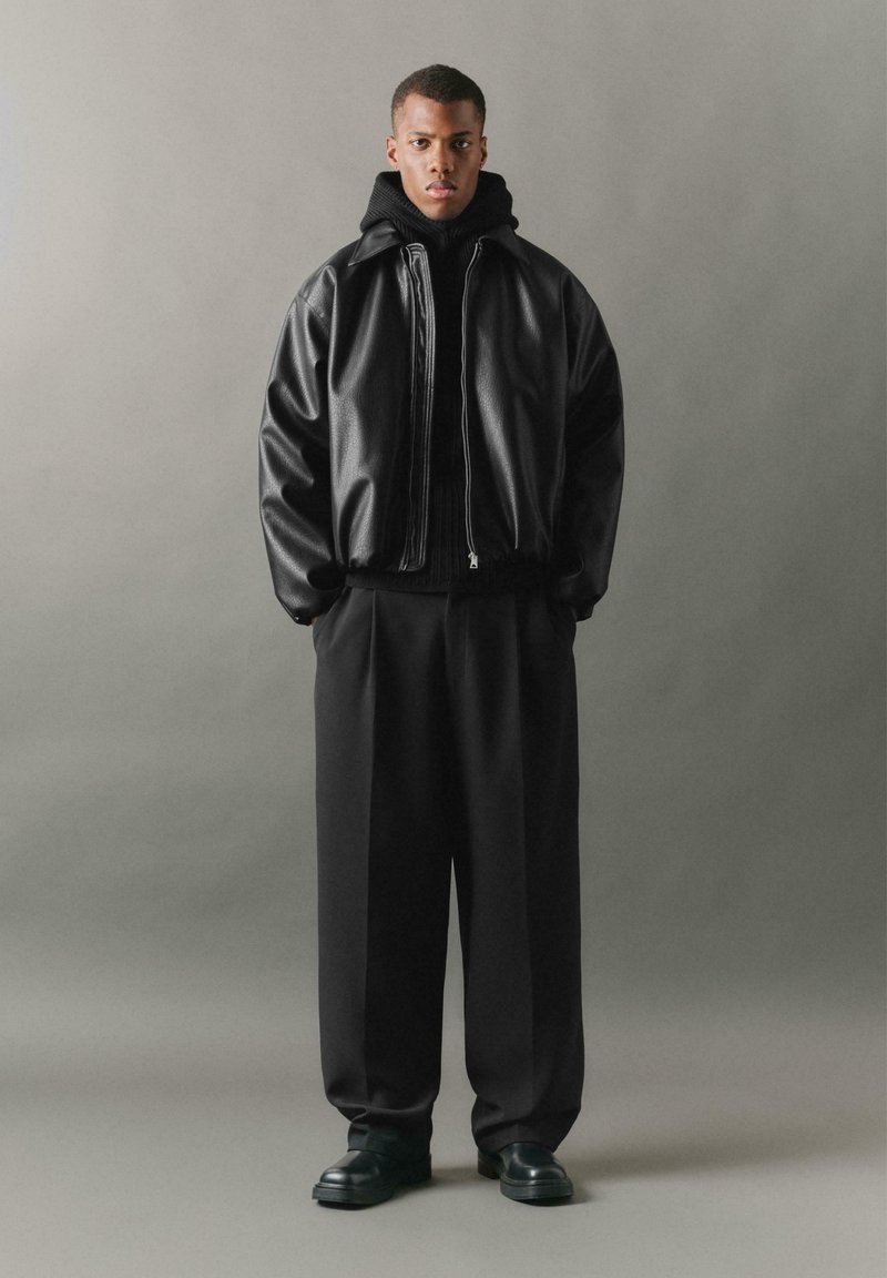 Giubbotto bomber in pelle nera con zip, colletto alto, abbinato a un maglione in maglia nera e pantaloni neri larghi. Scarpe nere spesse.