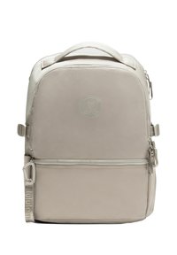 Béžový batoh Lululemon s horným uchom, prednou zipsovou kapsou, nastaviteľnými bočnými popruhmi a embosovaným logom na prednom paneli.