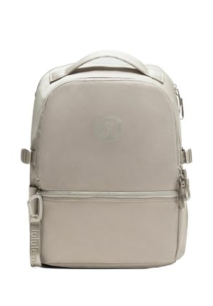 Beige Lululemon rugzak met bovenhandvat, voorzak met rits, verstelbare zijbanden en een reliëflogo op het voorpaneel.