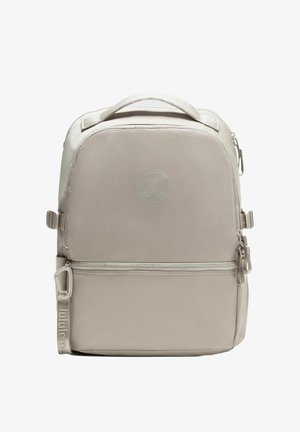 Beige Lululemon rugzak met bovenhandvat, voorzak met rits, verstelbare zijbanden en een reliëflogo op het voorpaneel.