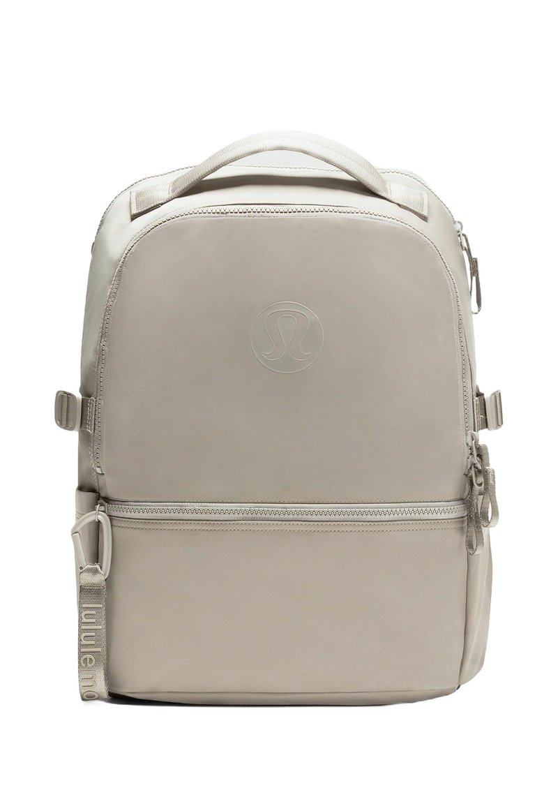 Béžový batoh Lululemon s horným uchom, prednou zipsovou kapsou, nastaviteľnými bočnými popruhmi a embosovaným logom na prednom paneli.