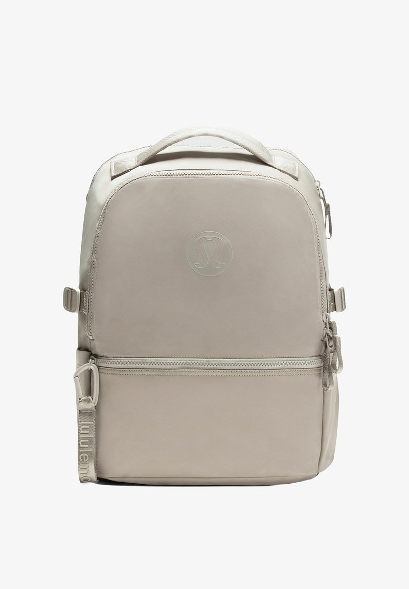 Beige Lululemon seljakott, millel on ülemine kätetoe, eesmine lukuga tasku, reguleeritavad küljetuged ja esipaneelil sisse pressitud logo.