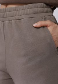 Pruun puuvillased dressipüksid elastse vöökohaga, küljetaskutega ja siledapinnalised, käsi taskus.