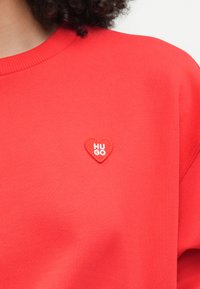 Κόκκινο φούτερ από μαλακό ύφασμα, με ένα μικρό κεντημένο πα patch σε σχήμα καρδιάς που γράφει "HUGO" στα λευκά. Ριμπέ σχέδιο στον λαιμό, άνετη εφαρμογή.