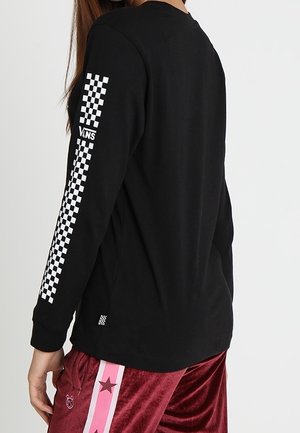 Persona con una camiseta negra de manga larga con el logo de Vans en tablero de ajedrez blanco en la manga y pantalones cortos rojos con una franja de estrella rosa.