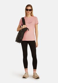 Rosa bomullströja med grafiskt tryck, svart axelväska, svarta leggings och beige sneakers. Solglasögon fulländar outfiten.