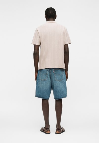 Άνδρας στέκεται με την πλάτη, φοράει μπεζ κοντομάνικη μπλούζα, μπλε τζιν σορτς και καφέ σανδάλια σε λευκό φόντο.