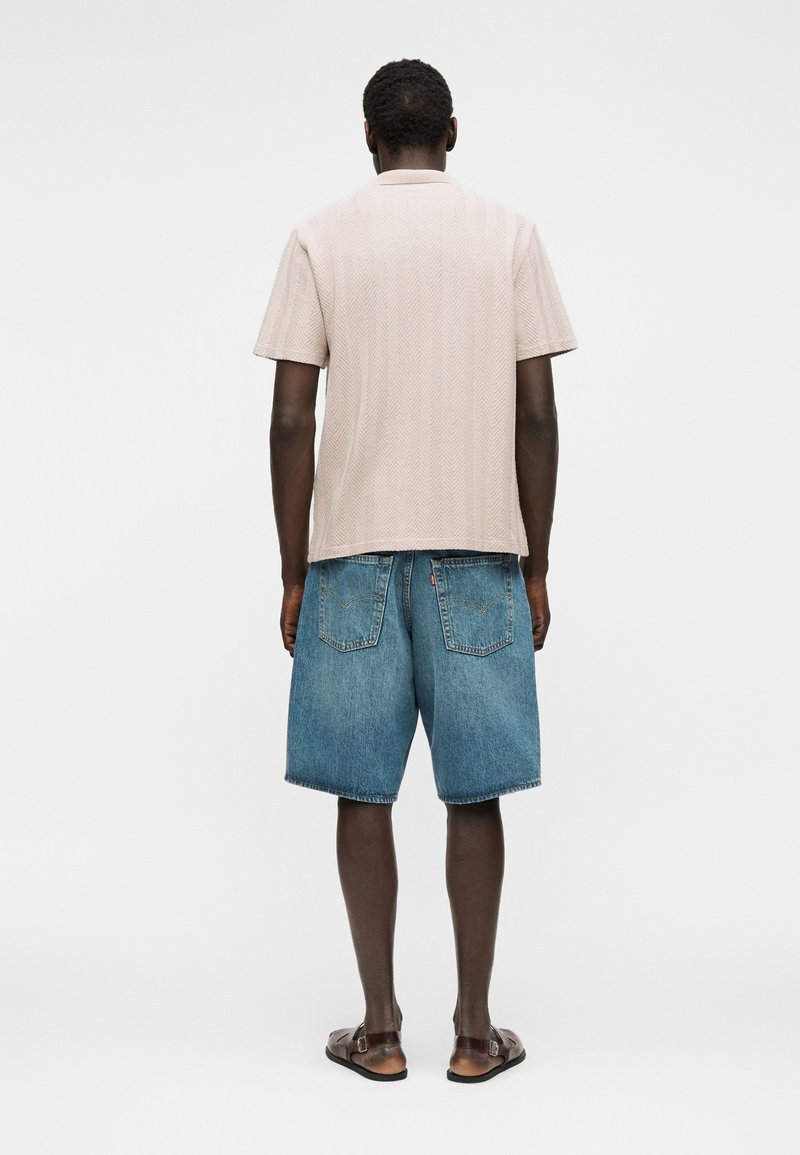 Un homme debout de dos, portant une chemise beige à manches courtes, un short en jean bleu et des sandales marron sur un fond blanc uni.