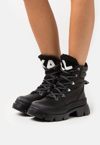 Botas de senderismo negras con parte superior de cuero texturizado, suela gruesa de goma y puño forrado de forro polar con letras blancas.