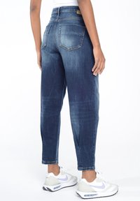 Mörkblå högmidjade jeans med något avsmalnande ben, två bakfickor och en urtvättad yta, tillsammans med vita och grå sneakers.