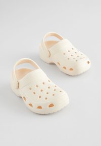 Világos bézs klumpák rugalmas műanyagból, több szellőzőnyílással a légáteresztésért, hátsó pánttal és texturált talppal.