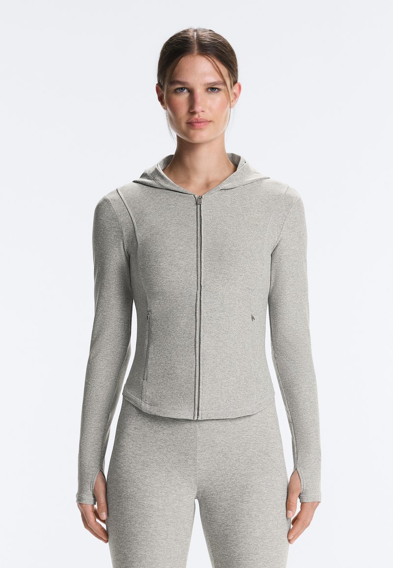Femme portant un sweat à capuche zippé gris clair avec des trous pour les pouces et un pantalon ajusté assorti, debout devant un fond clair uni.