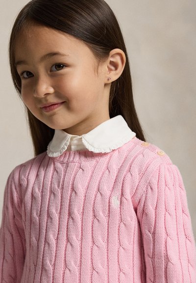 Jeune fille aux longs cheveux foncés portant une chemise blanche à col et un pull rose en maille torsadée, souriant légèrement et regardant la caméra.
