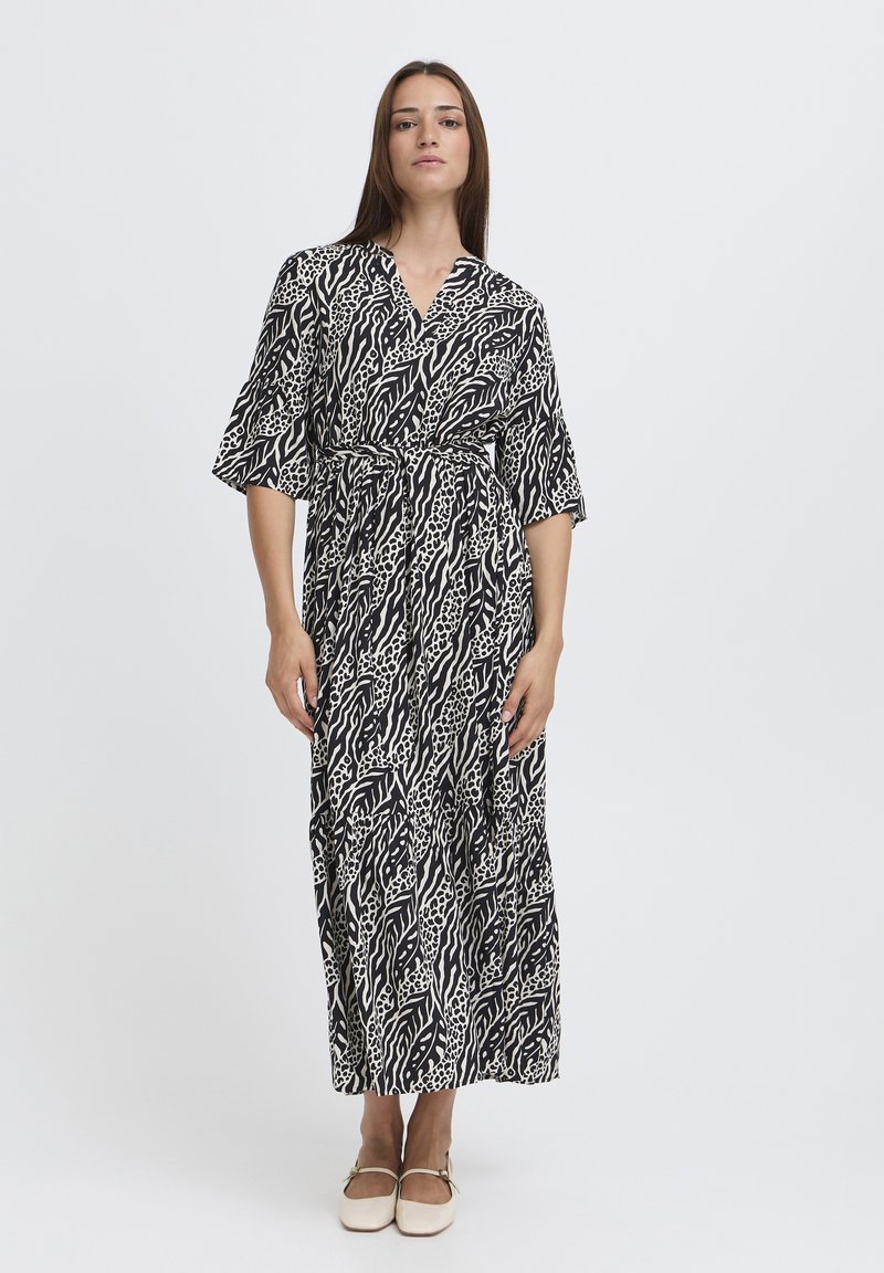 Femme debout portant une robe midi ceinturée à imprimé animalier noir et blanc avec des manches longueur coude et des chaussures Mary Jane crème sur fond blanc.