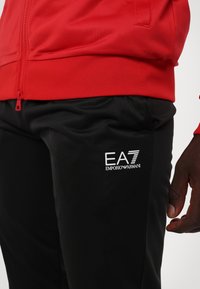 Sarkans rāvējslēdzēja jaka ar teksturētu virsmu, apvienota ar melniem sporta biksēm, kuru sānos ir balts EA7 Emporio Armani logo.