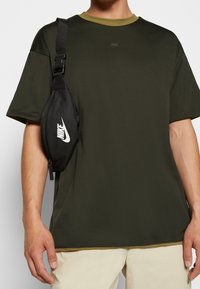 Riñonera negra de Nike con logo blanco, llevada sobre una camiseta de manga corta verde con un acabado texturizado y un cuello de un verde claro contrastante.