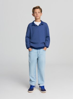 Maglione a maglia blu con colletto, abbinato a jeans chiari strappati. L'outfit è completato da sneakers navy con dettagli bianchi.