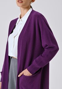 Lila gestrickte Strickjacke mit offenem Vorderteil, gerippten Kanten und Seitentaschen, getragen über einer weißen Bluse, kombiniert mit grauen Hosen.