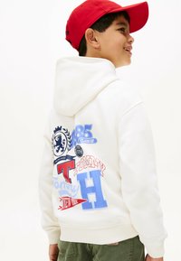 Felpa bianca con cappuccio regolabile, caratterizzata da grafiche stampate colorate sul retro, inclusi loghi e testo in rosso, blu e nero.