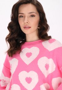 Rosa Strickpullover mit großen weißen Herzmustern, verziert mit kleinen Perlen, Rundhalsausschnitt und lockerer Passform. Glatte Textur.