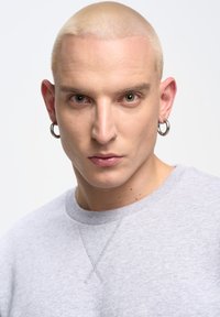 Šedá mikina s kulatým výstřihem, s viditelnými detaily šití. Osoba má krátké, platinově blond vlasy a stříbrné kruhové náušnice.