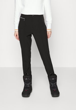 Personne portant un pantalon noir sur mesure avec poche zippée et des bottes de neige noires, debout devant un fond blanc uni.