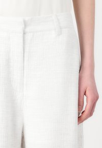 White textured pants with a crinkled fabric finish. Features a flat waistband and side pockets, partially visible hand on the right.